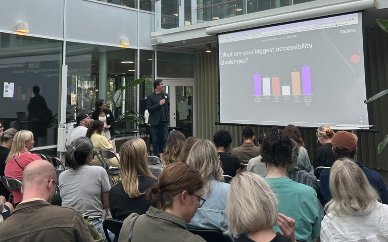 Chris stands in front of an audience of seated people, holding a microphone. On a screen is a slide reading 'What are your biggest accessibility challenges?', and the results of a Menti survey.