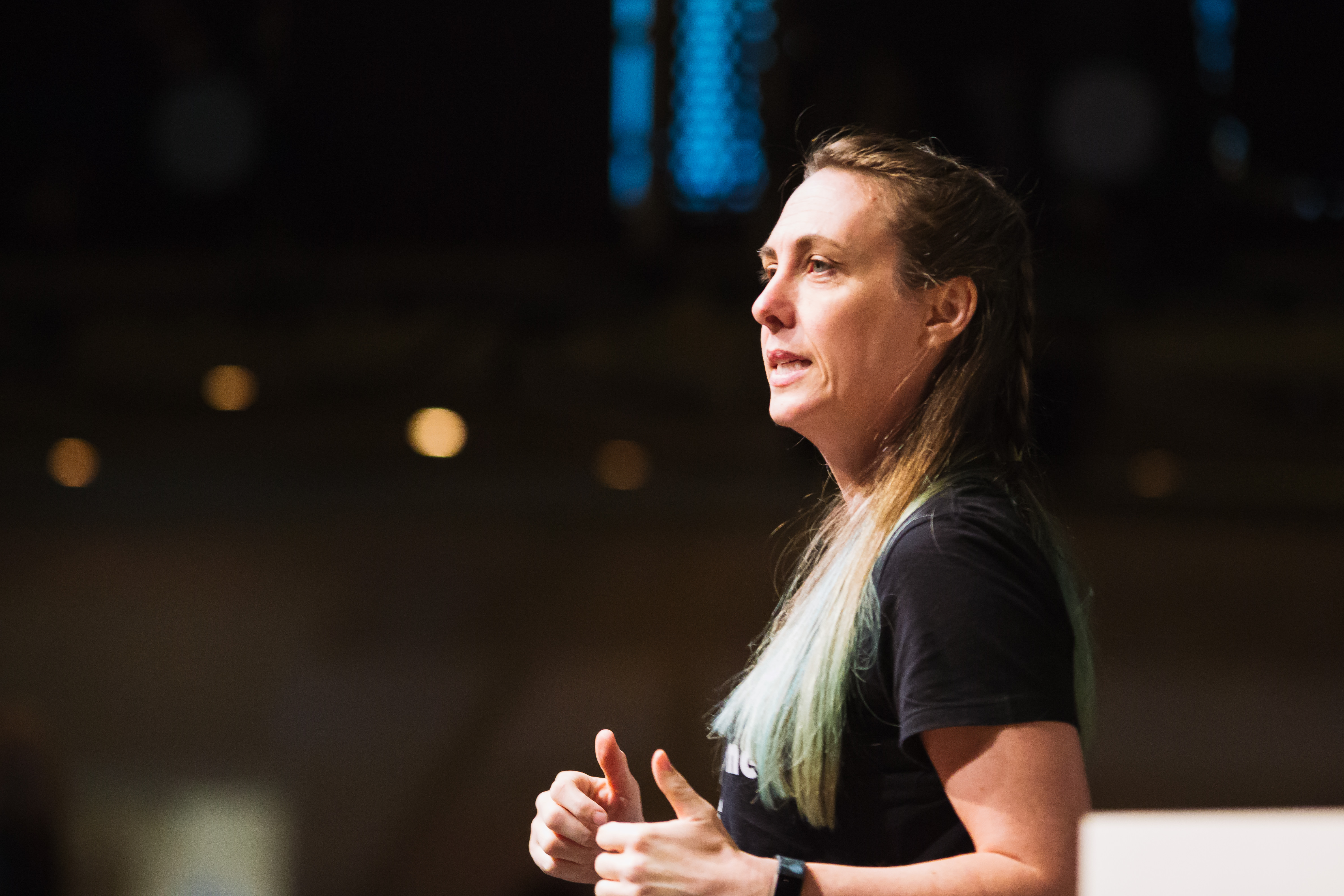 Sarah Richards. A white woman with long brown hair and turquoise dyed tips of her hair. She is in profile and her hands are raised with her thumbs up as she is gesticulating. 