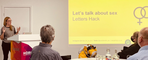Emma Parnell gives a talk to a conference room of seated people. Behind is a slide reading 'Let's talk about sex'