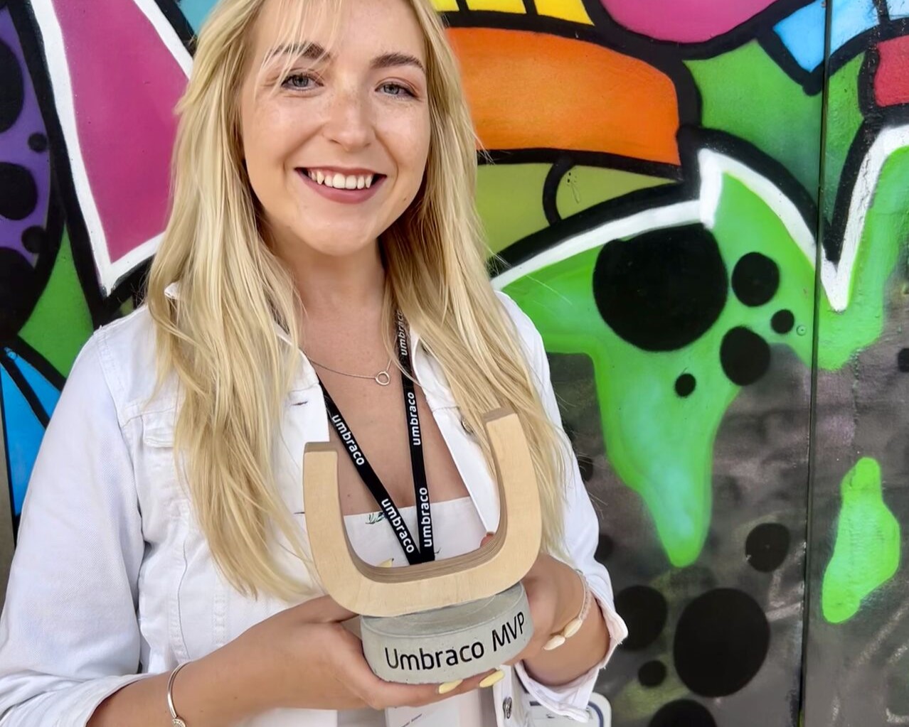Amy smiles at the camera, holding her U-shaped Umbraco MVP award. She has blonde hair and blue eyes, and is wearing a pale pink top and white denim jacket.