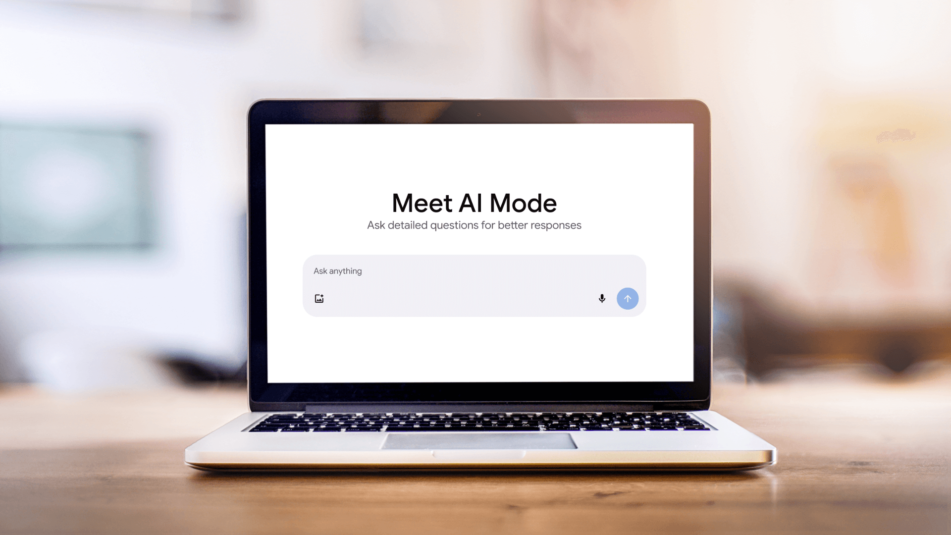 A laptop sits open on a wooden table indoors, displaying a screen with the heading “Meet AI Mode.” Below the heading is a prompt box that reads “Ask anything.” The room in the background is softly blurred, keeping the focus on the laptop’s screen.