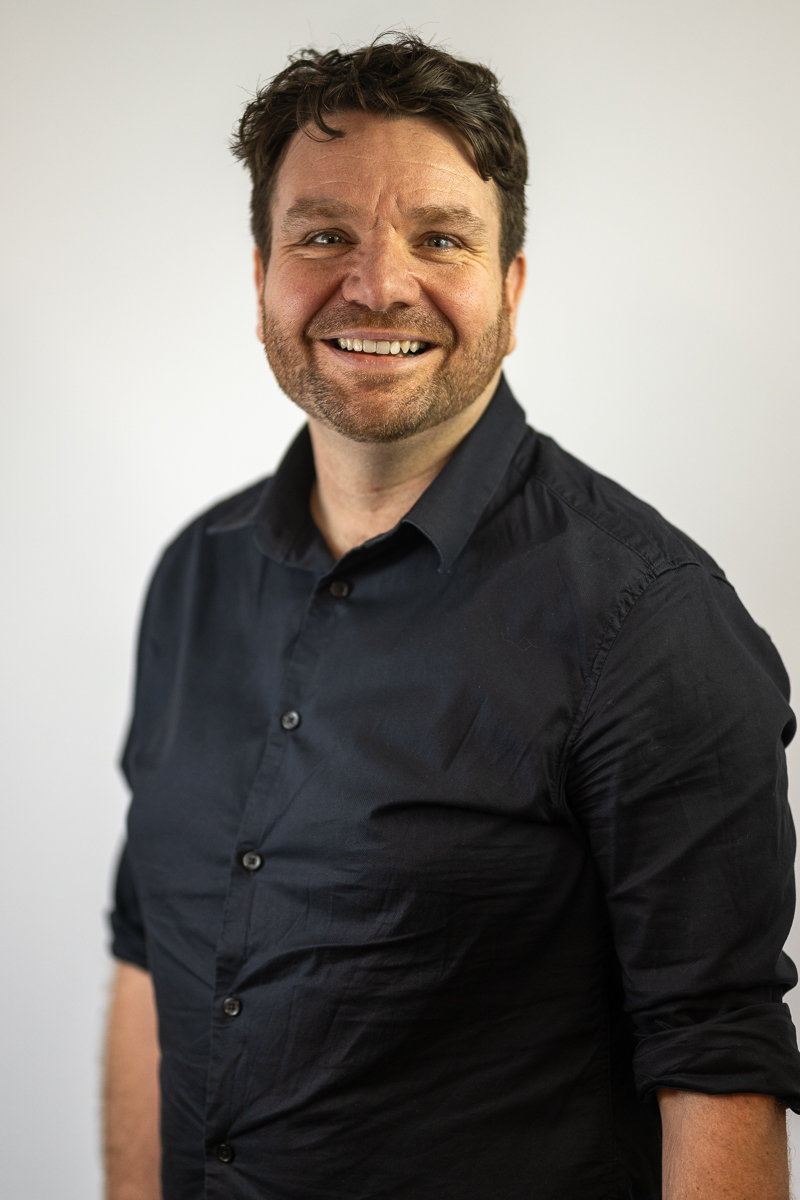 Hugh is standing against a plain white background and smiling. He has short dark hair and a beard, wearing a dark button-up shirt