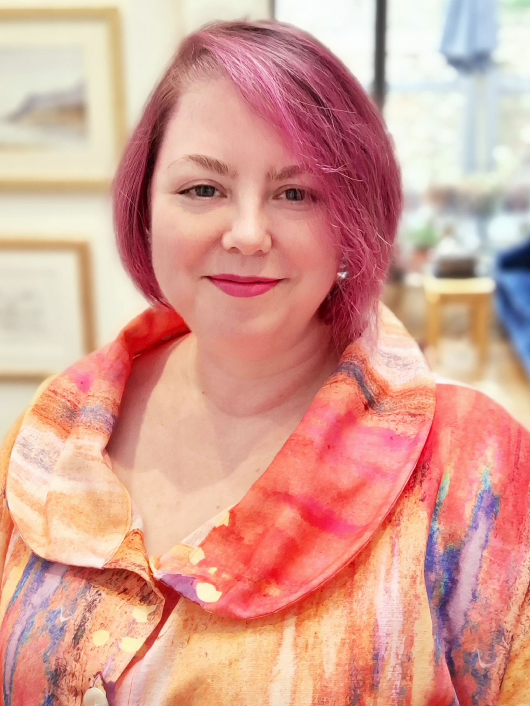 Claire is smiling at the camera. Her hair is a deep pink colour, and she is wearing a pink and orange shirt with a watercolour-esque design.