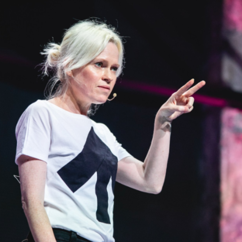 Tash Willcocks speaking on stage, wearing a white T-shirt and gesturing with one hand during a presentation.