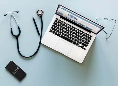 An open laptop on a table with a mobile phone, glasses, a pen and a stethoscope laying around it