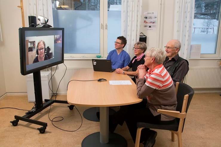 Three older white people with a white nurse sat around a table looking at a large television on a mobile stand. They are on a video conference and a white lady's face is displayed on the tv screen. 