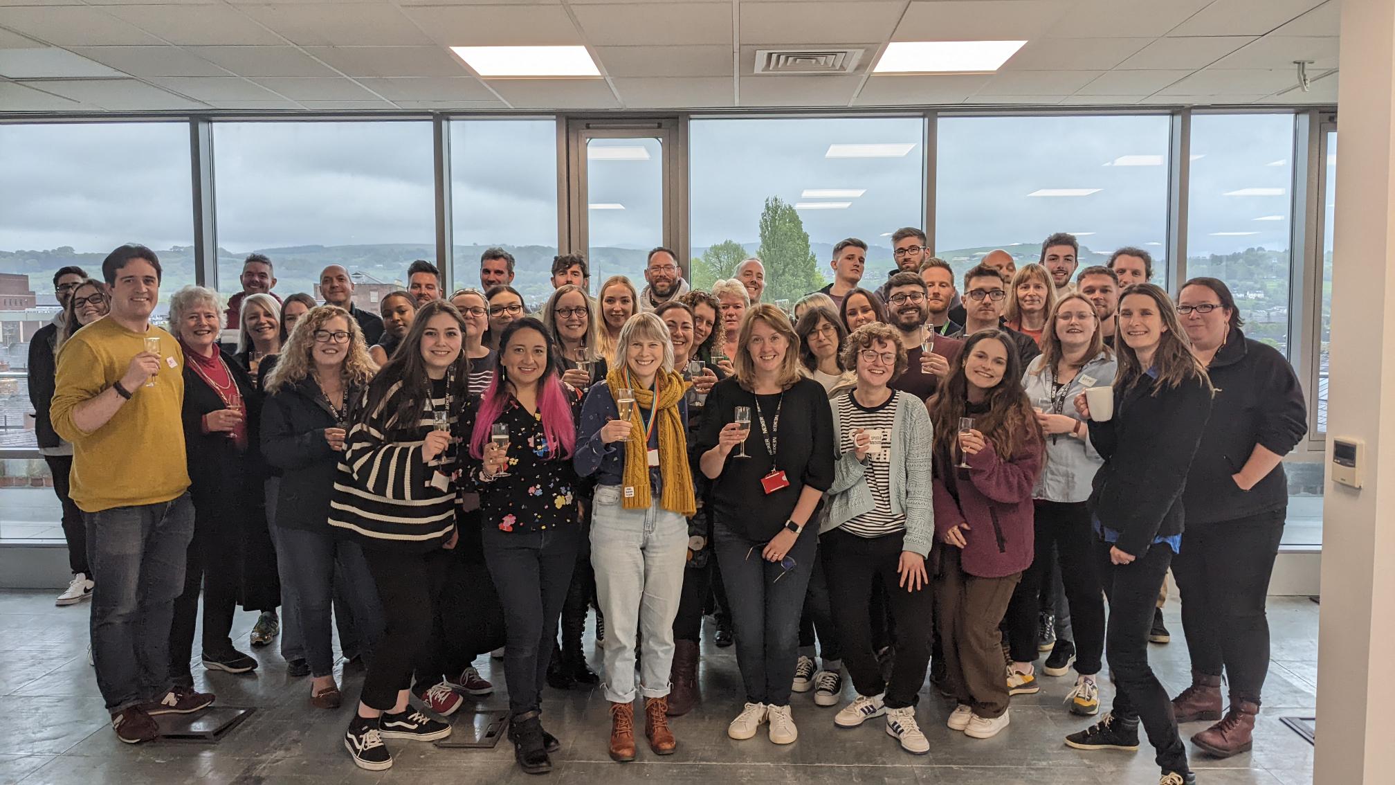 A group of about 65 people standing in an office with green hills in the background.