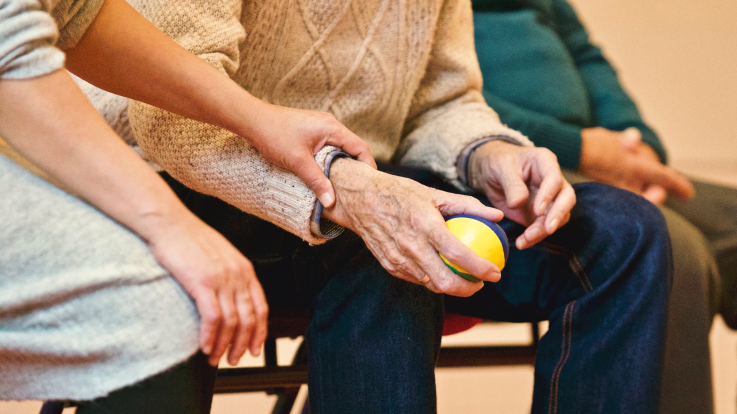 An elderly hand holding a ball. Another hand is resting on this hand with the ball. 