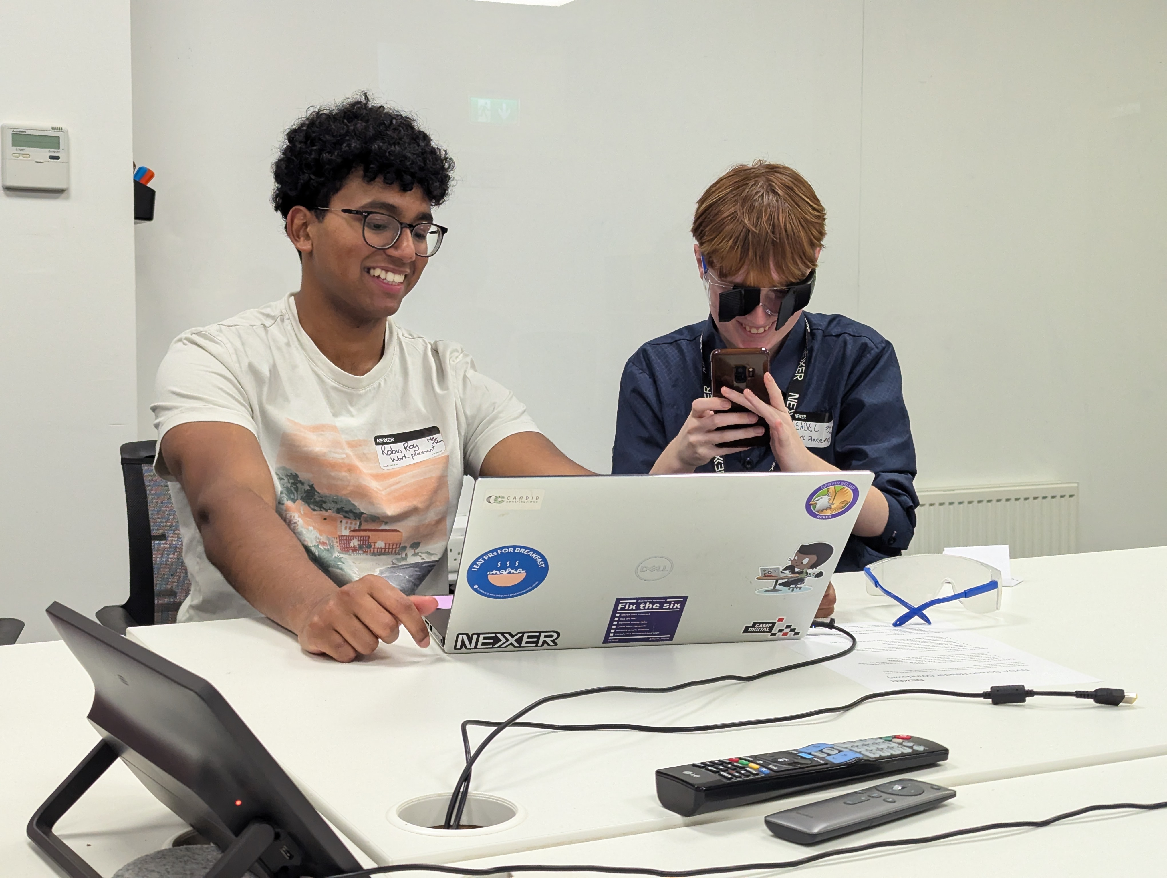 Robin and Isobel using the popup accessibility lab. Robin has brown eyes and hair, and is smiling while using a laptop. Isobel has brown hair, and is wearing simulation goggles. On the table is another pair of simulation goggles. 
