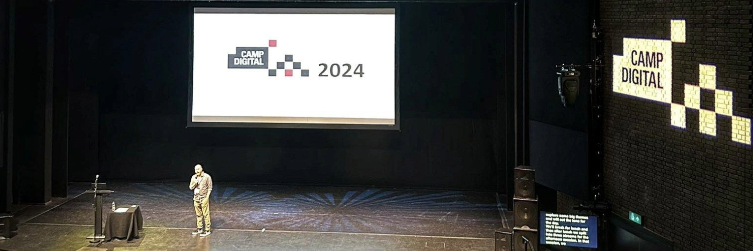Camp Digital host Shaun Gomm stands onstage at the RNCM, with the Camp Digital logo visible behind and on the wall