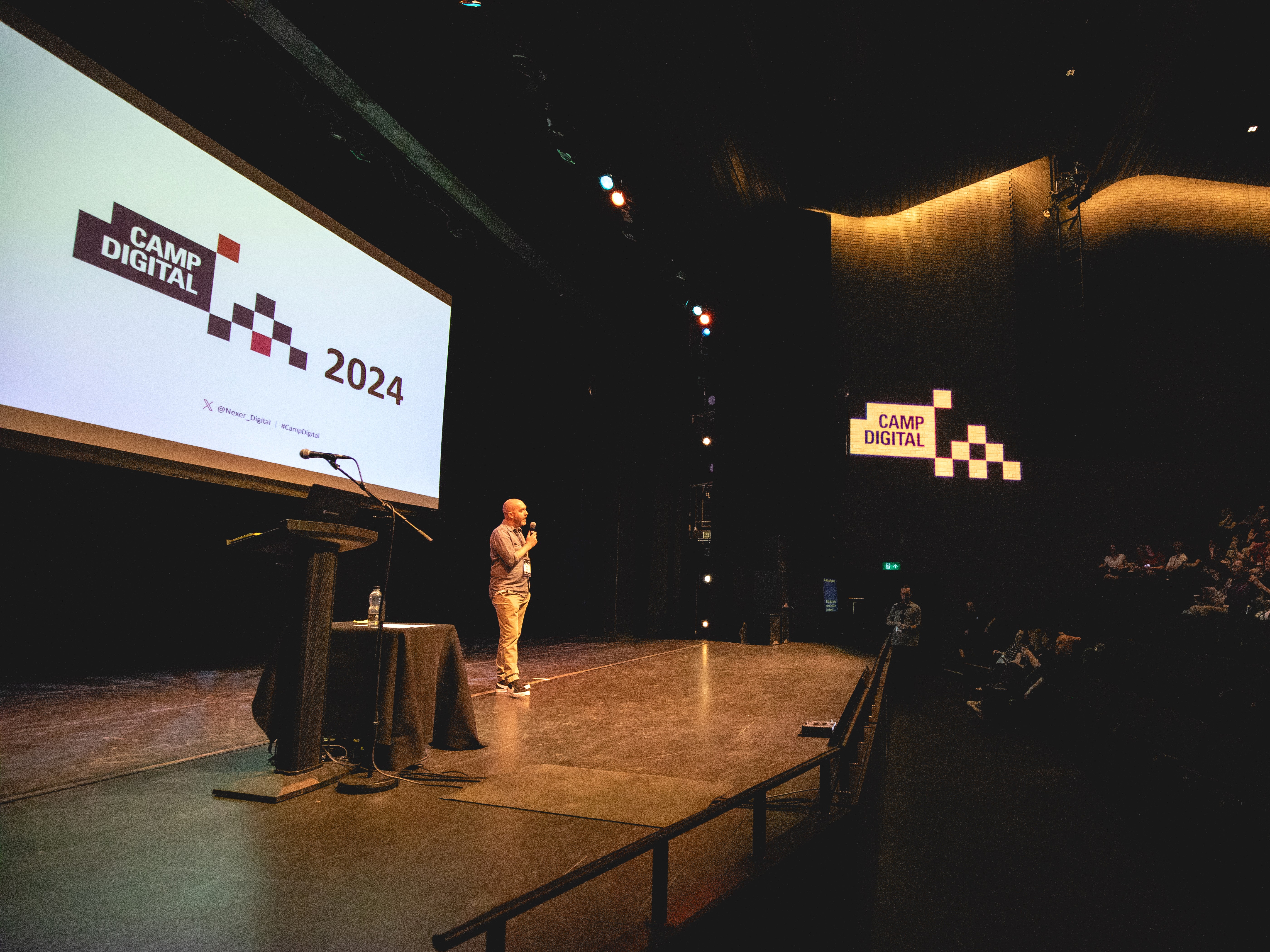 Camp Digital host Shaun Gomm stands onstage at the RNCM, with the Camp Digital logo visible behind and on the wall