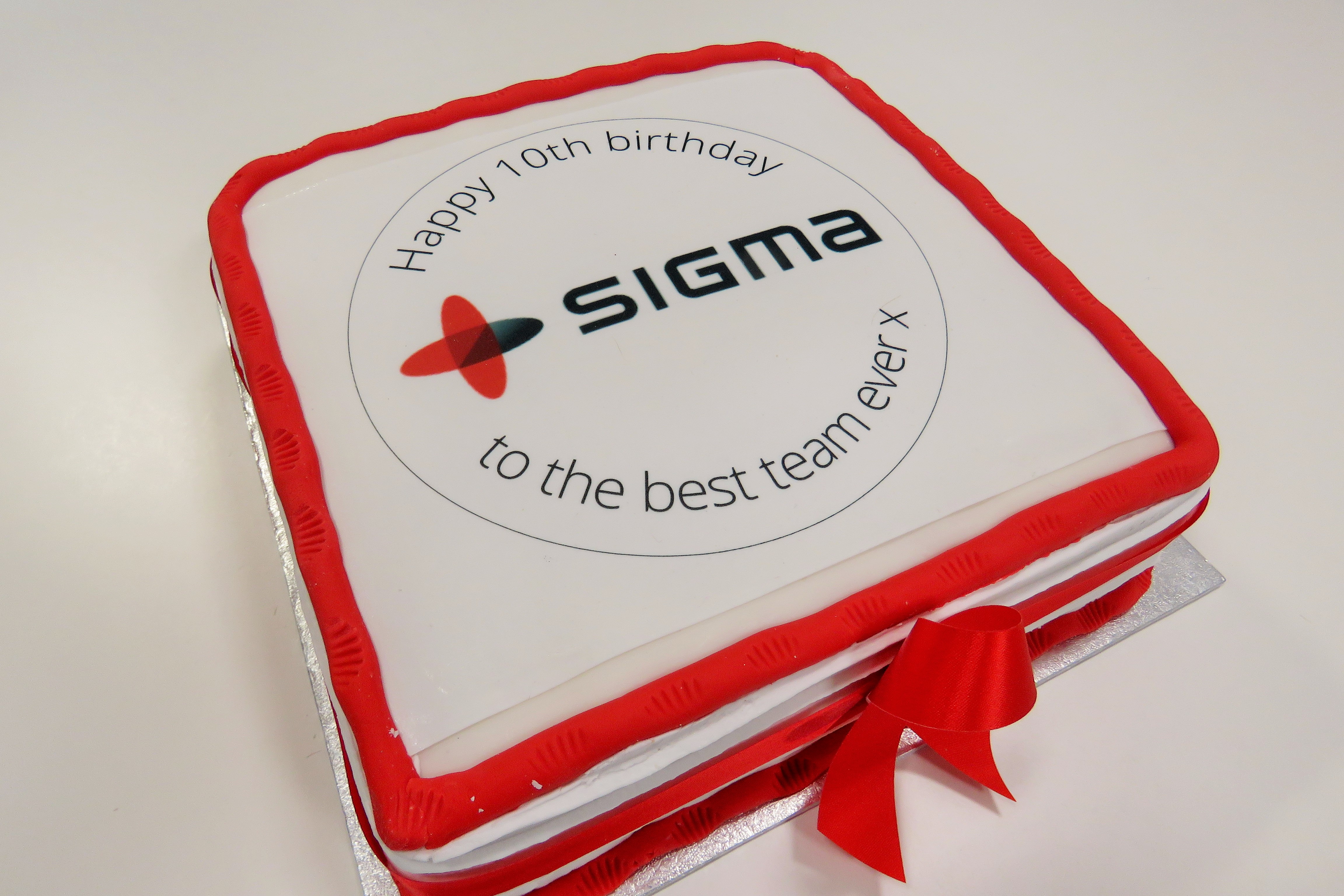 A white cake with an iced red border with the Sigma logo on it and words reading, "Happy 10th birthday to the best team ever x"