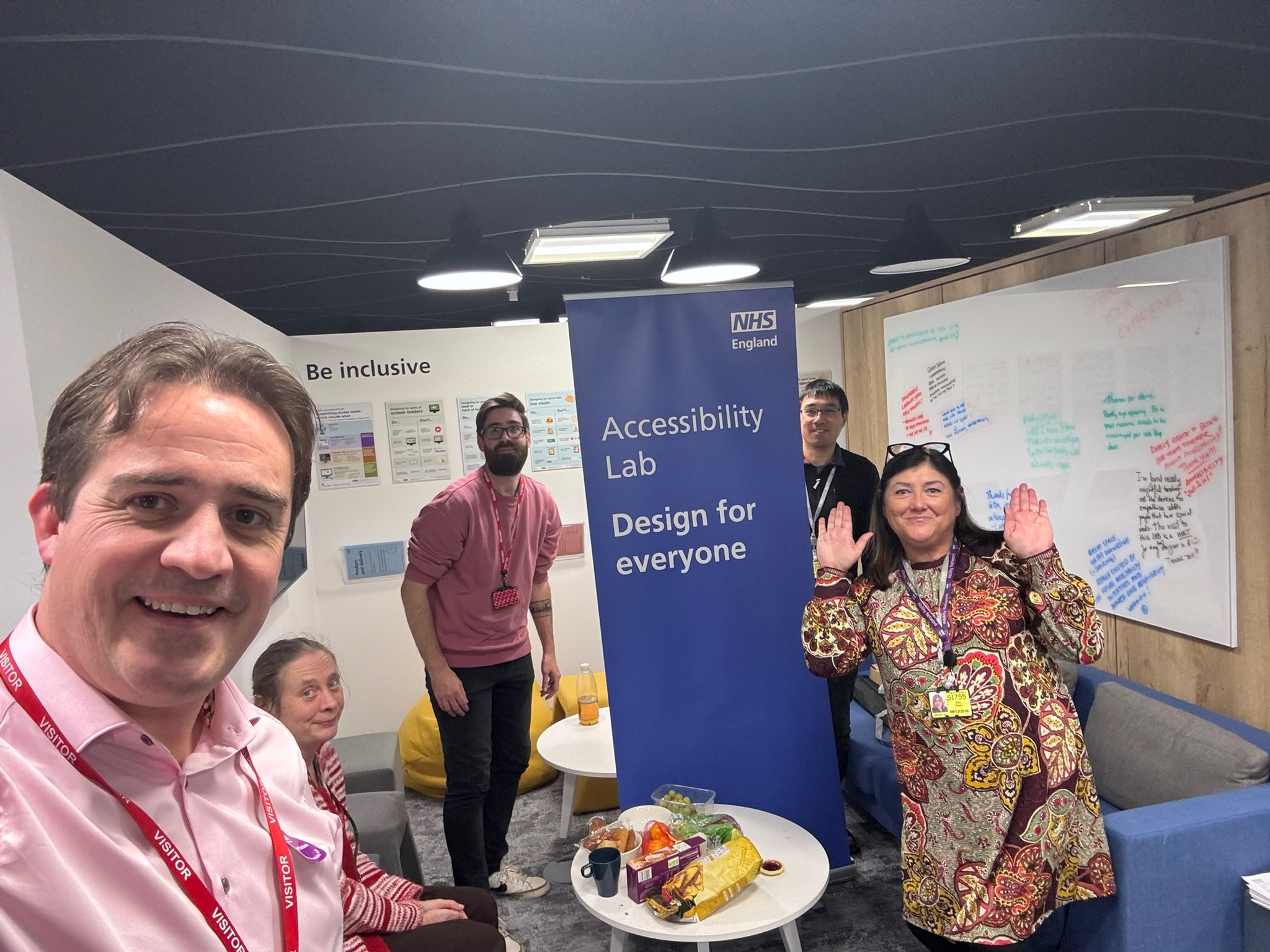 Chris and Danny smile at the camera, along with others on the team who worked on the NHSE safety lab. Chris is wearing a pink shirt, and has brown eyes and hair. Danny is wearing a pink jumper and has brown eyes and hair. A roll up banner next to Danny reads "NHS Accessibility Lab, Design for everyone"