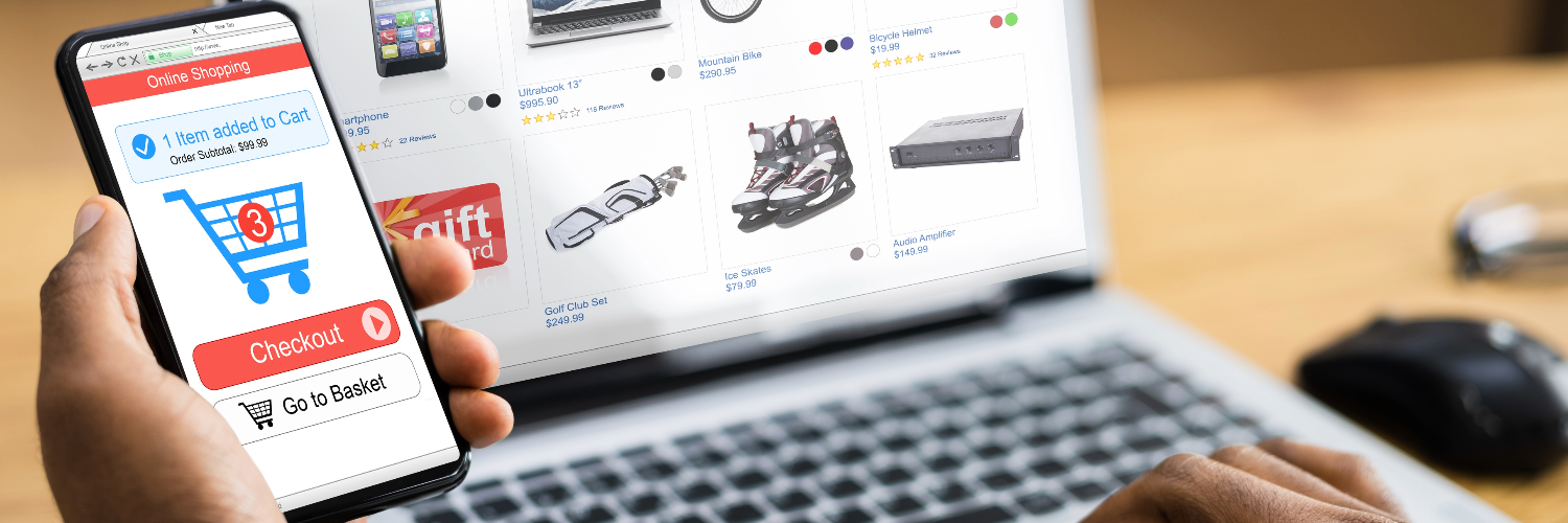 A person holds a phone in one hand, with a checkout basket displayed on the screen. On a laptop screen, being typed by the other hand, is an eCommerce interface