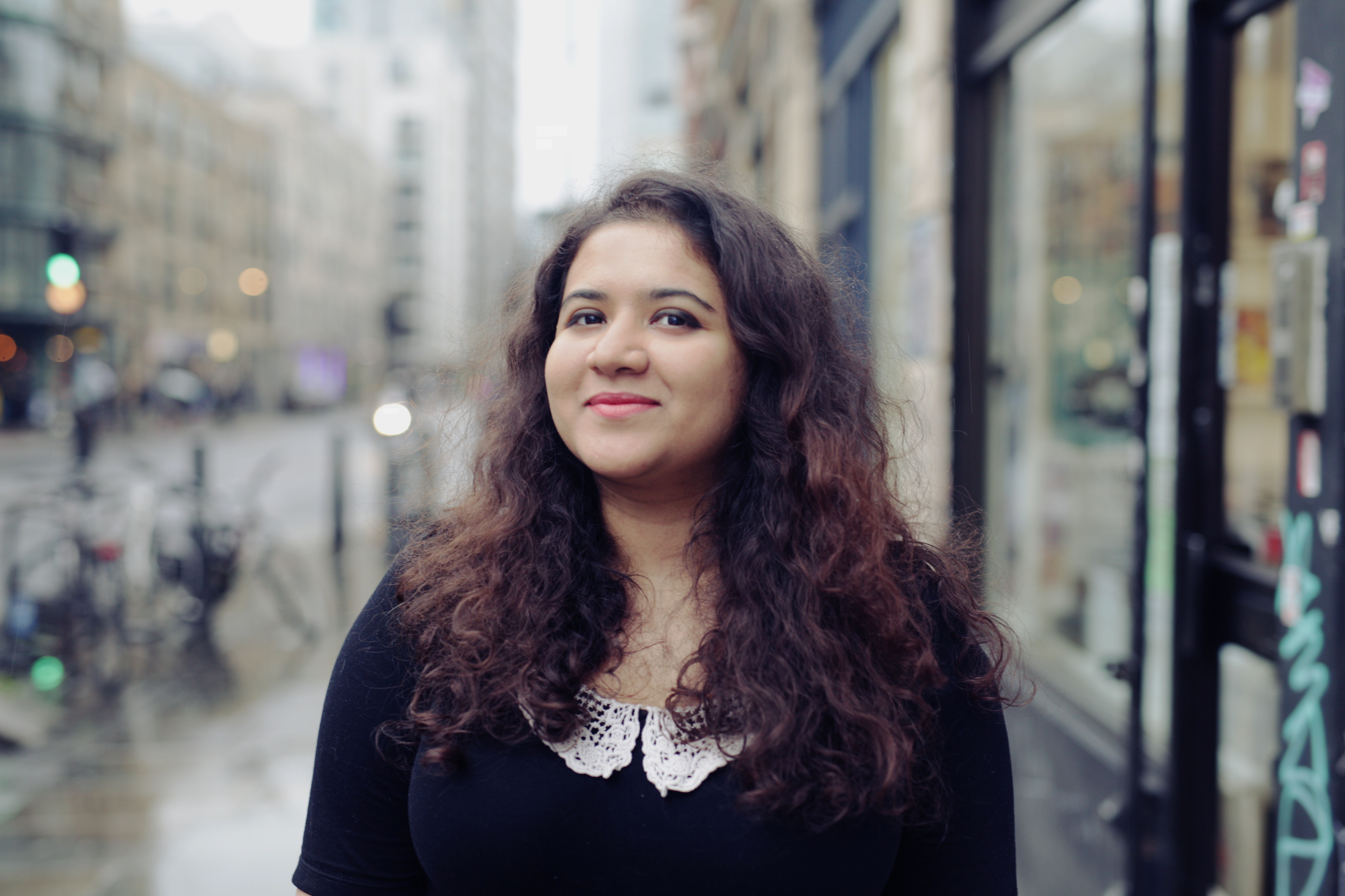 Hera grins at the camera. She has long, curly hair. Stands on a city street, wearing a black top with a white lace collar. The background is blurred, showing buildings and parked bicycles on a rainy day
