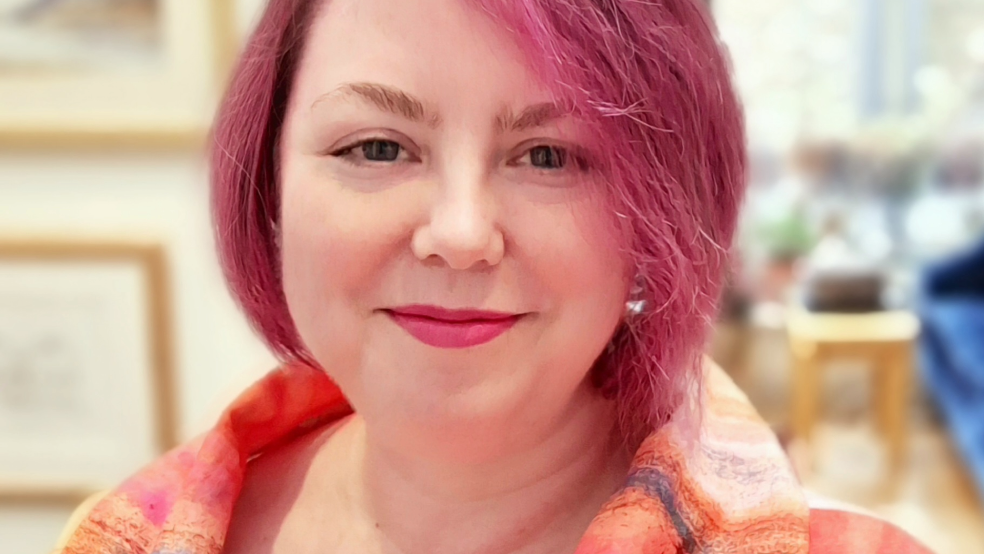Claire is smiling at the camera. Her hair is a deep pink colour, and she is wearing a pink and orange shirt with a watercolour-esque design.