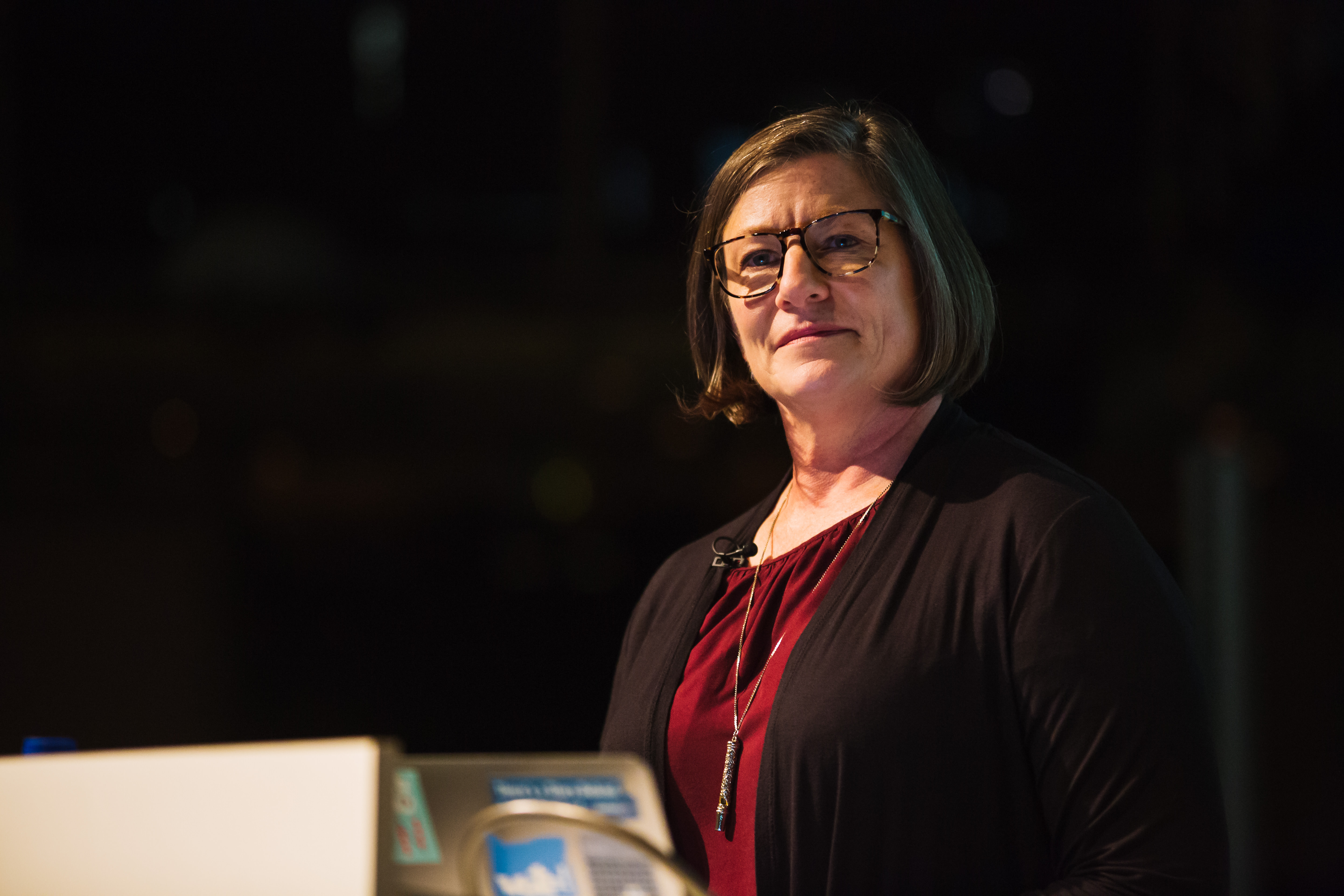 Dana Chisnell. A white woman with short brown hair and glasses. She is wearing a necklace and a maroon top. Her head is tilted to the side and she looking thoughtful.