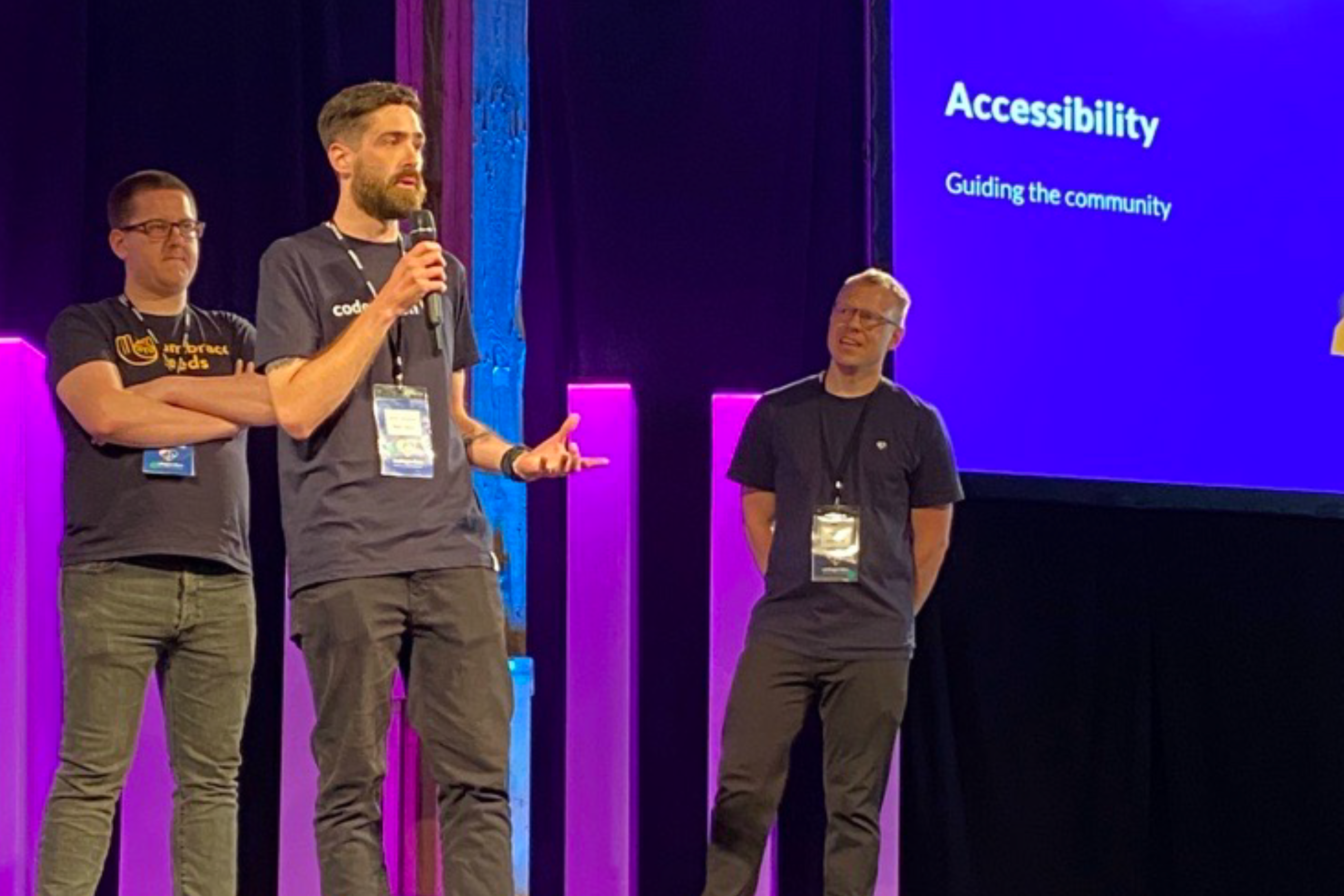Three people standing on stage with a presentation behind them reading 'accessibility, guiding the community'