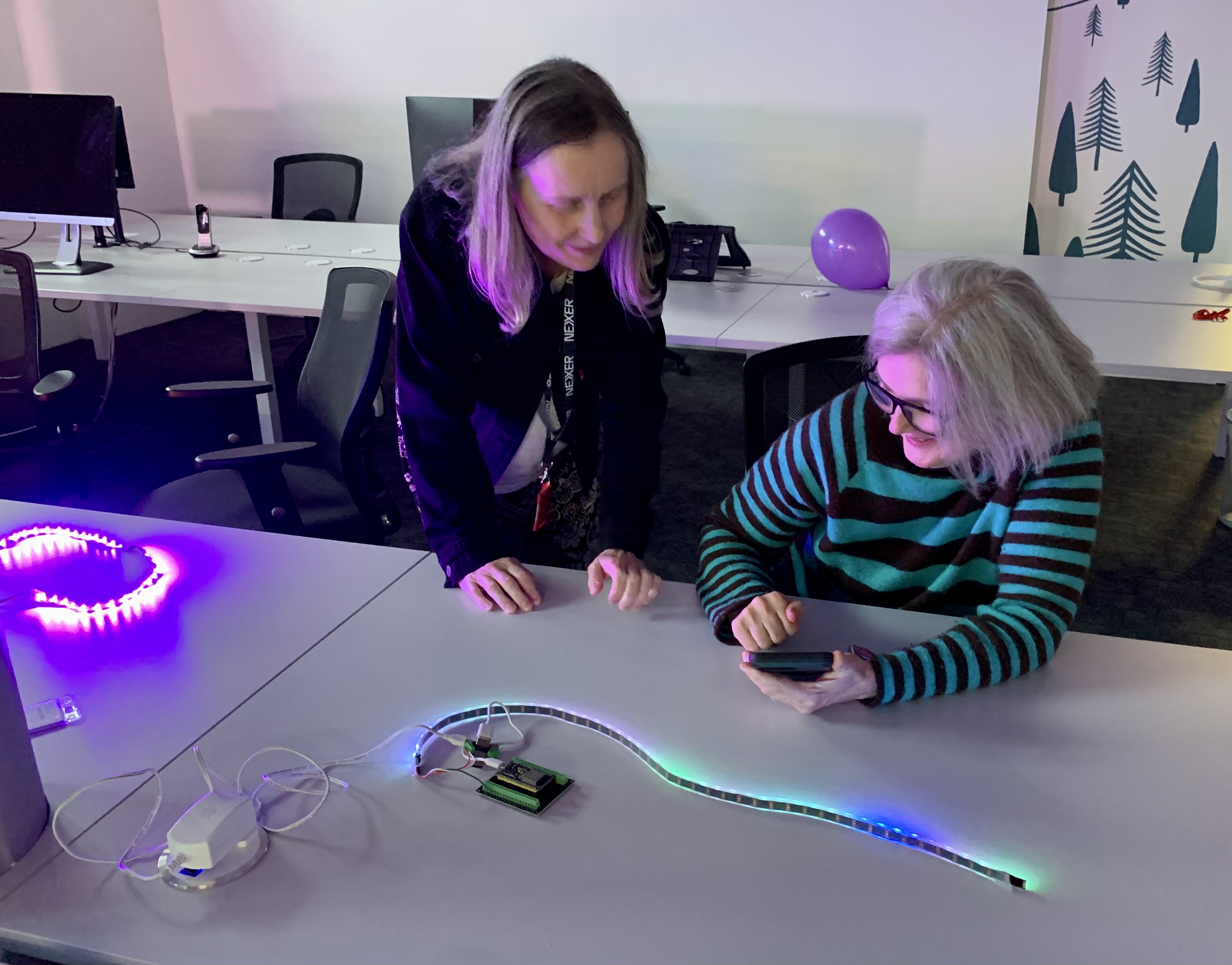 Two women, Cat and Mel, at a desk with blue and green light coming from strips on the desk. Both women are looking at a phone and smiling.