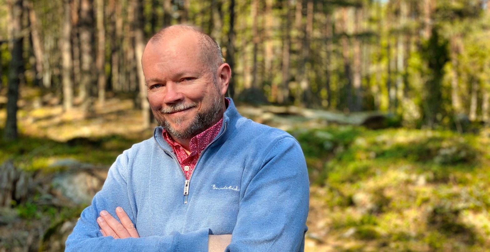 Per is crossing his arms while standing happily in a sunlit forest. He is wearing a light blue sweater over a red patterned shirt. The background features tall trees and greenery