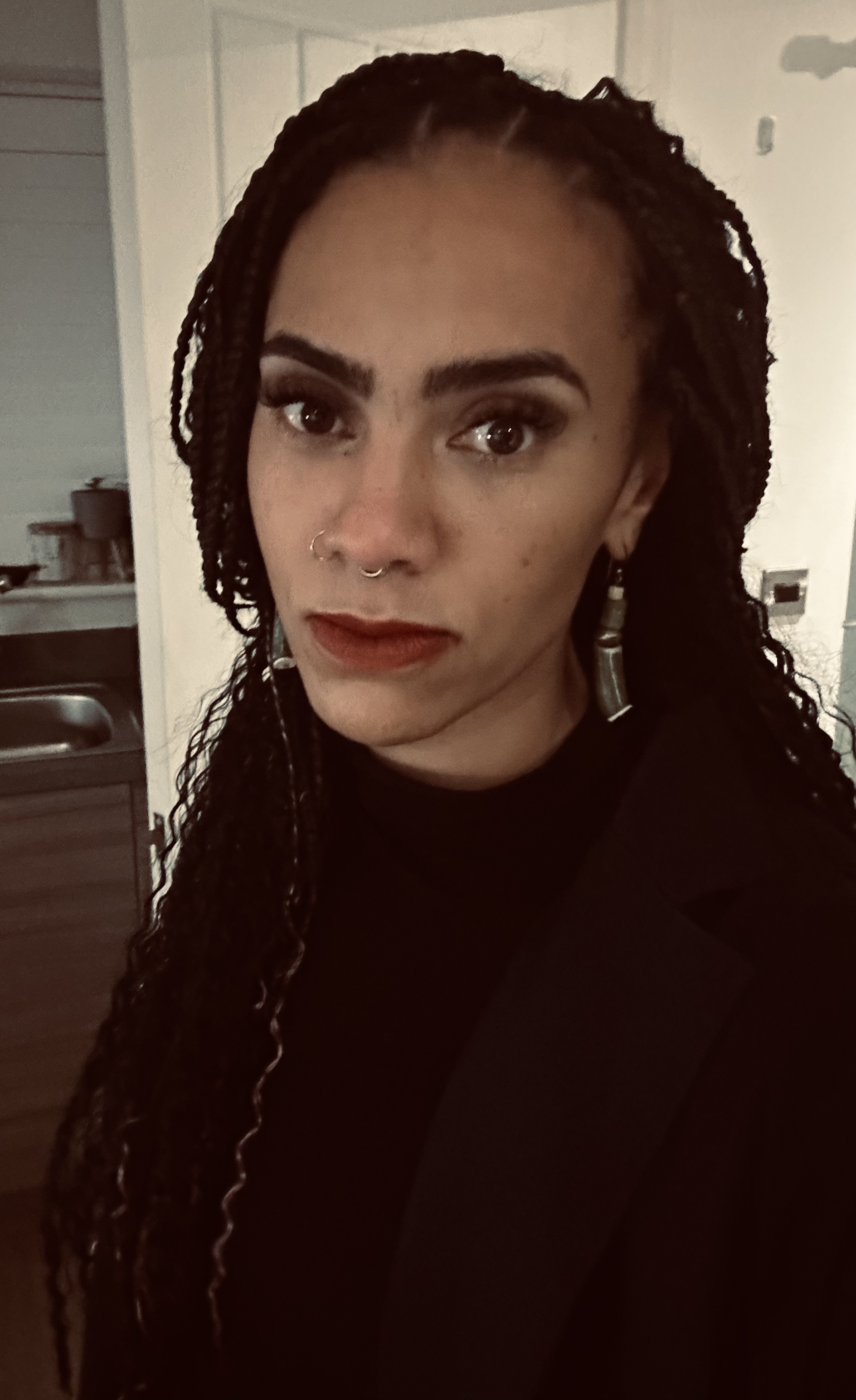 Kanika has braided hair and a nose ring stands in a room with soft lighting. She is wearing a dark top and earrings. They have a confident expression and are looking directly at the camera.